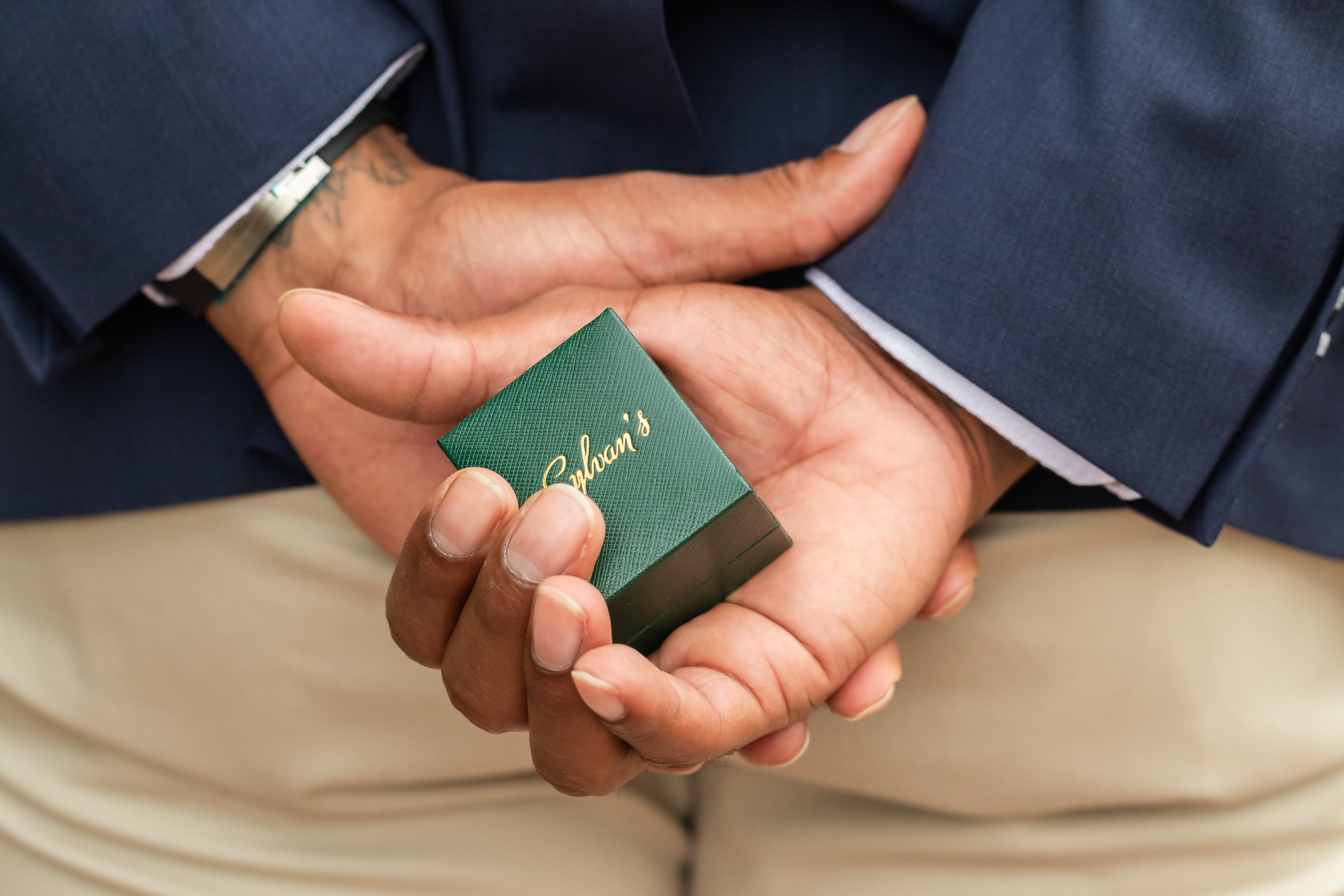 A man dressed elegantly, holding a ring box behind his back, symbolizing a romantic surprise or proposal. The setting conveys anticipation and a heartfelt moment.